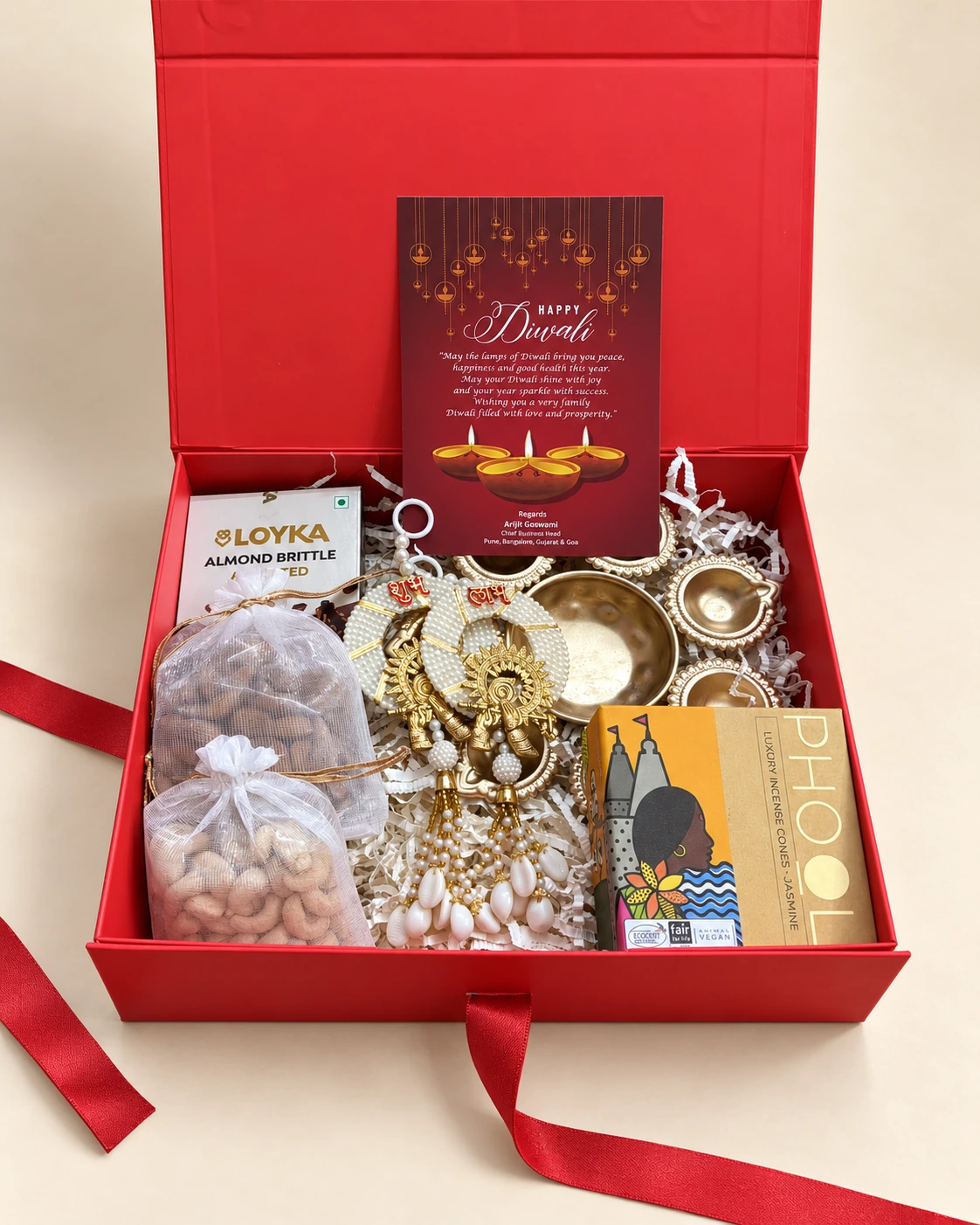 Open red Diwali hamper with dry fruits, almond brittle, brass diyas and a greeting card