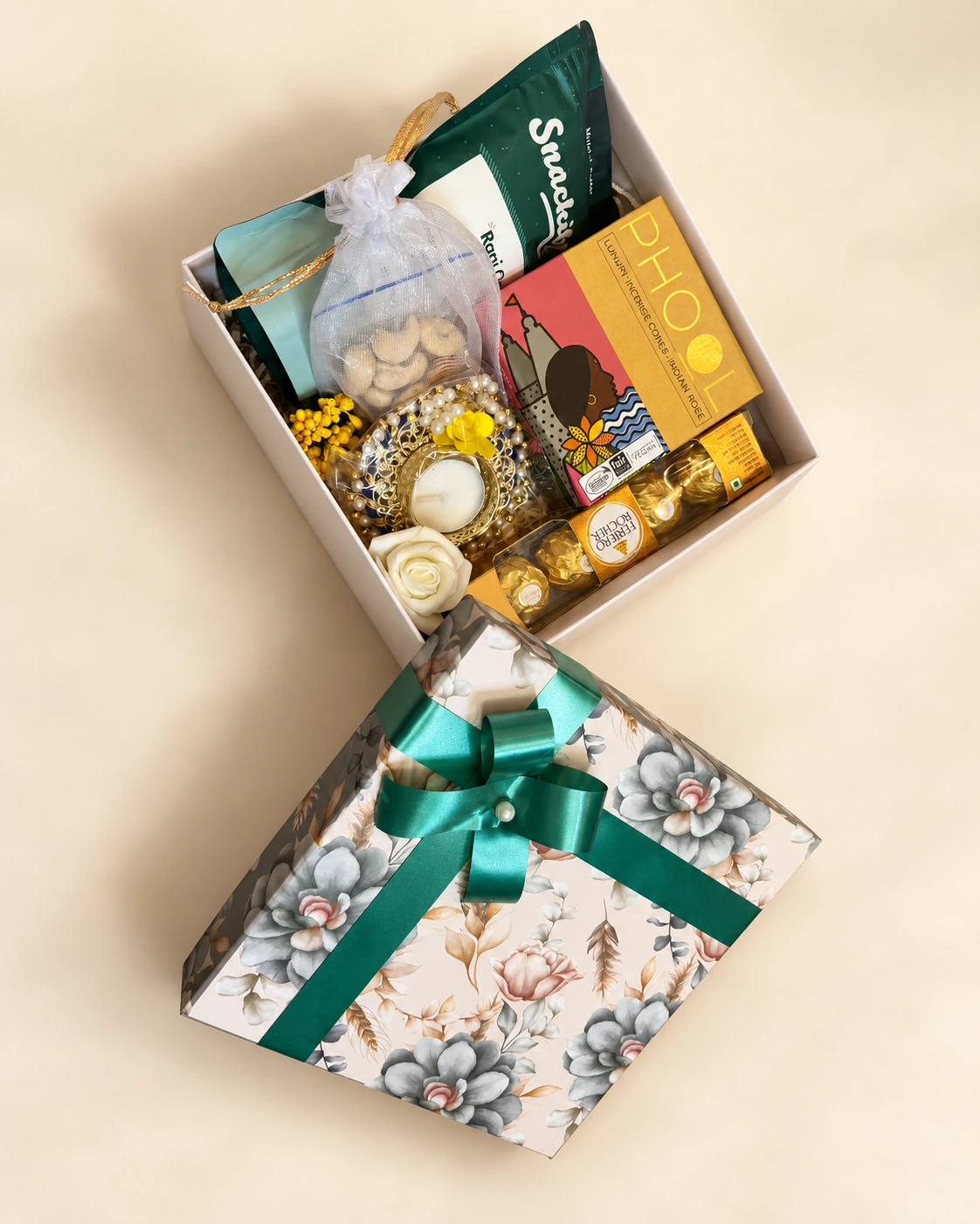 Open gift box with snacks and a candle beside a floral lid tied with a green ribbon